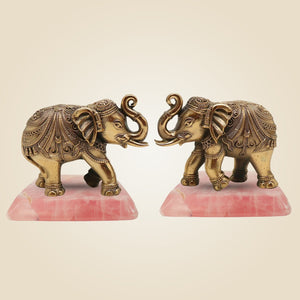 Two ornate brass elephant statues on pink stone bases against a beige background